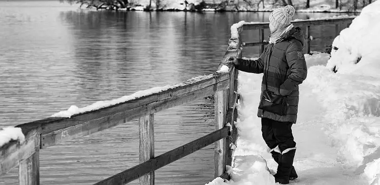 Eine Frau steht im Winter auf einem schneebedeckten Weg und schaut auf einen See. Symbolbild für den Text, der erklärt, ab welcher Temperatur und wie viel Grad unsere Atemluft sichtbar ist.